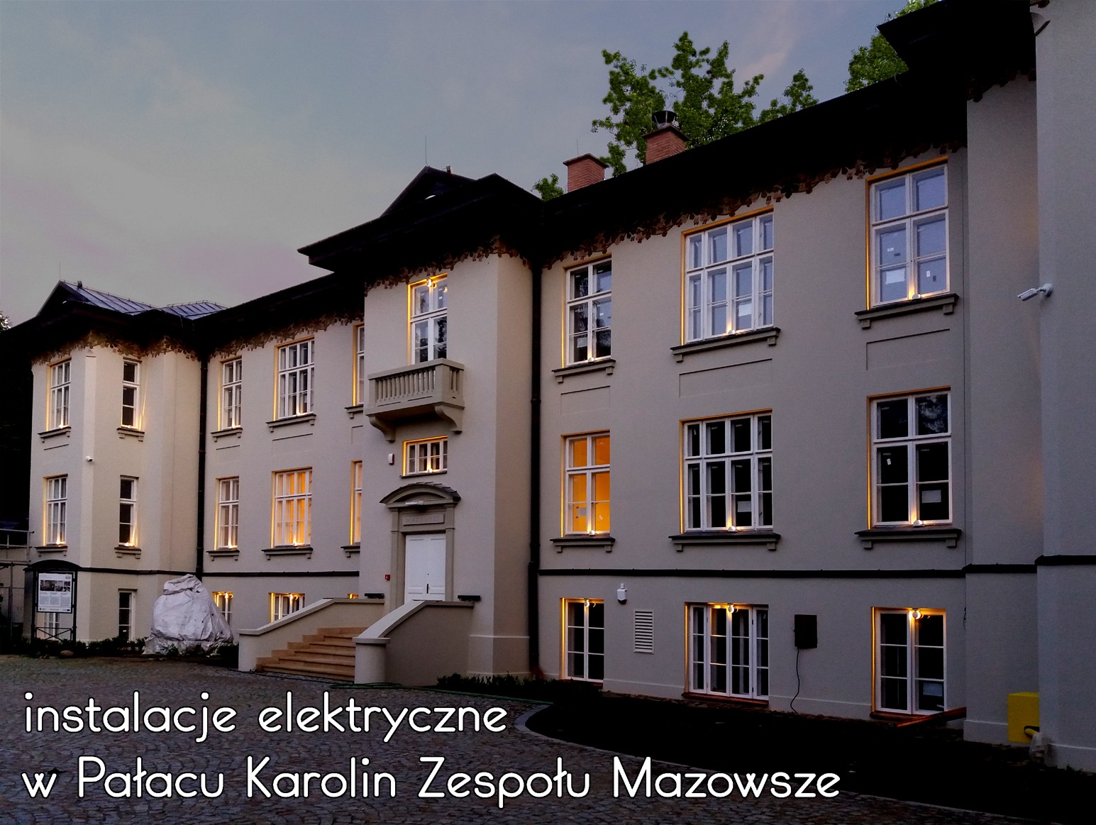 Pałac Karolin Zespołu Mazowsze oświetlony po zmroku, widoczne lampy elewacyjne wokół okien i drzwi, kamera monitoringu na ścianie budynku.