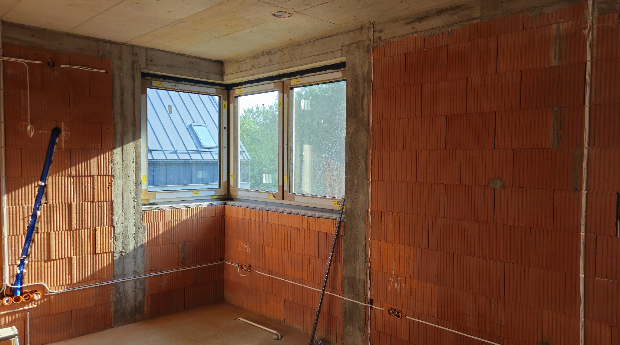 Surowy stan wnętrza z oknami i instalacją elektryczną w korygowanej ścianie z cegły. Widoczne kable i rury instalacyjne. Częściowo zabetonowany strop.
