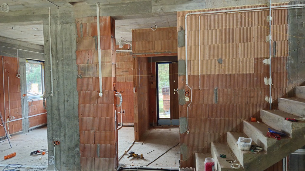 Wnętrze w trakcie prac instalacyjnych: widoczne kable elektryczne prowadzone po ścianach z cegły, surowy beton, narzędzia na schodach, okno z widokiem na zieleń.