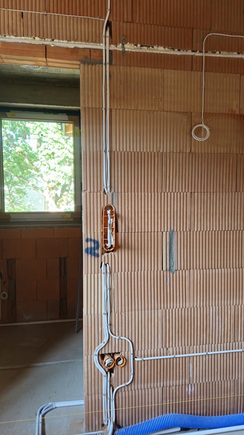 Instalacja elektryczna w surowym domu: kable w ścianie z cegły, puszki instalacyjne. Widoczne okno z zielenią na zewnątrz. Niebieska rura karbowana na dole.