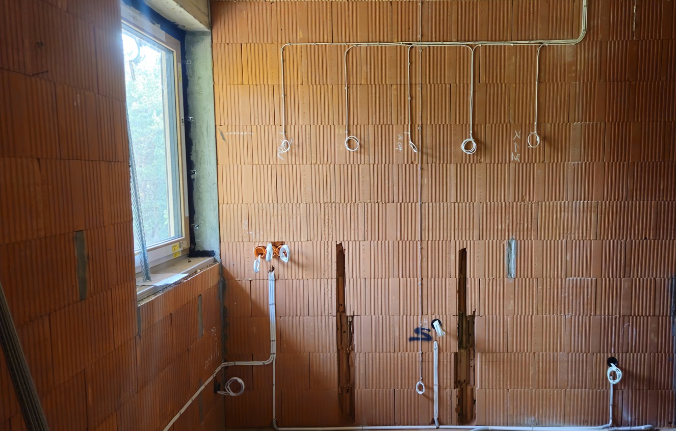 Wewnętrzna instalacja elektryczna w budynku w trakcie budowy. Widoczne przewody w ścianach z cegły ceramicznej, gniazdka i puszki. Okno z widokiem na zewnątrz.