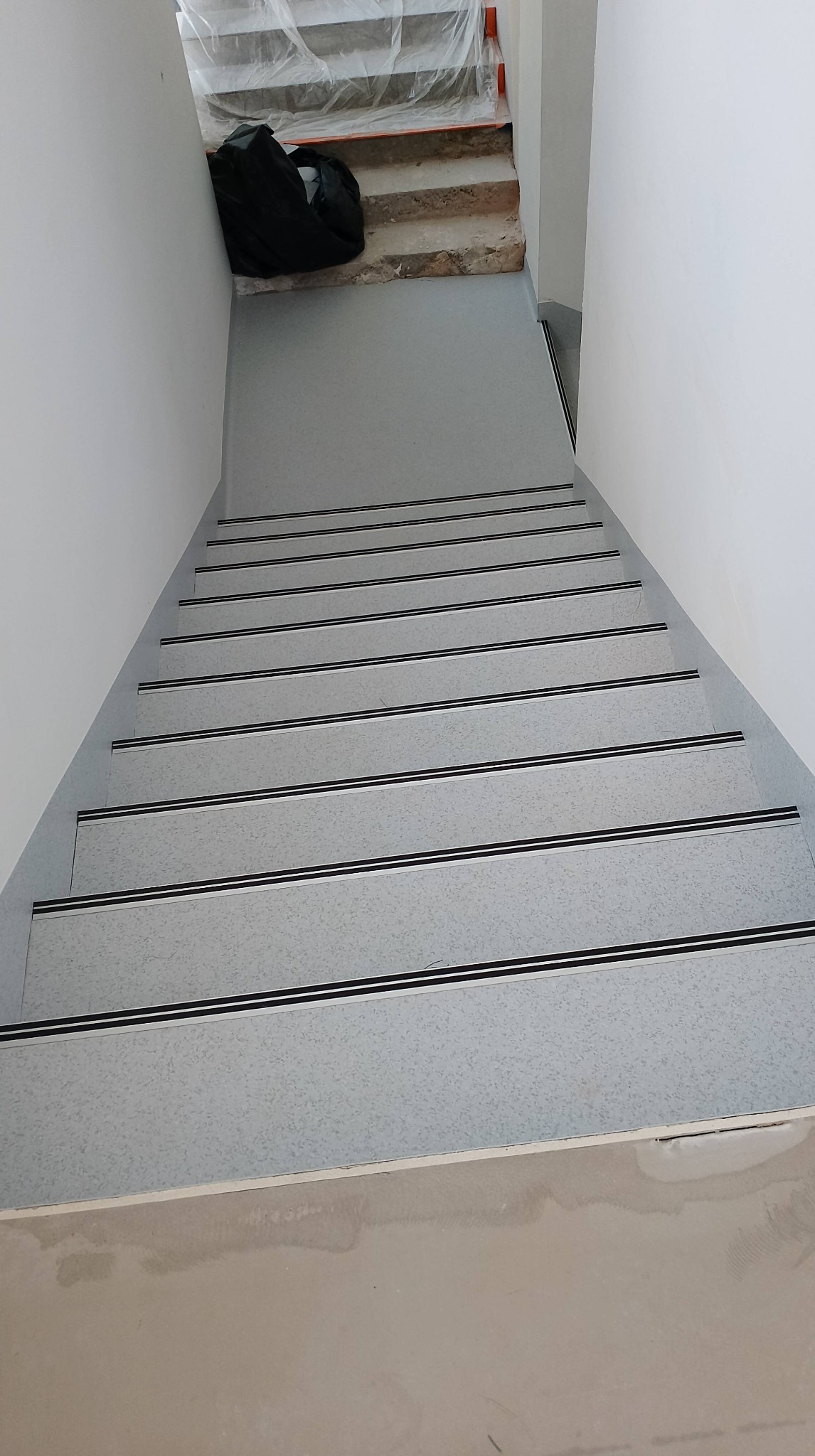 Schody pokryte jasnoszarą wykładziną z czarnymi paskami antypoślizgowymi, widok z góry, z workiem budowlanym na górnym podeście i fragmentem schodów w trakcie remontu.