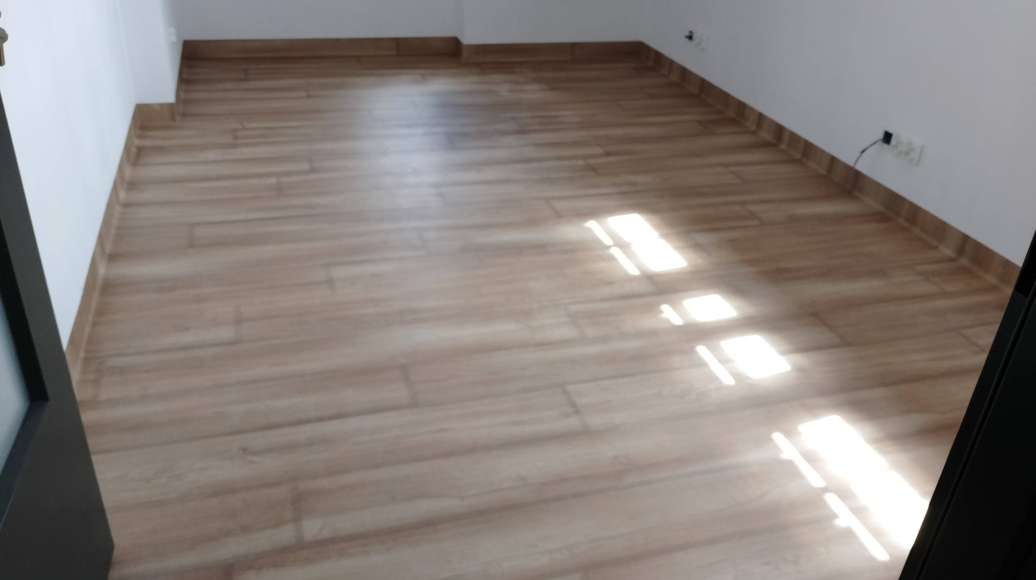 Jasne panele podłogowe imitujące drewno, ułożone w nowym pomieszczeniu z białymi ścianami i widocznym gniazdkiem elektrycznym, fragment czarnej ramy drzwi po lewej stronie.