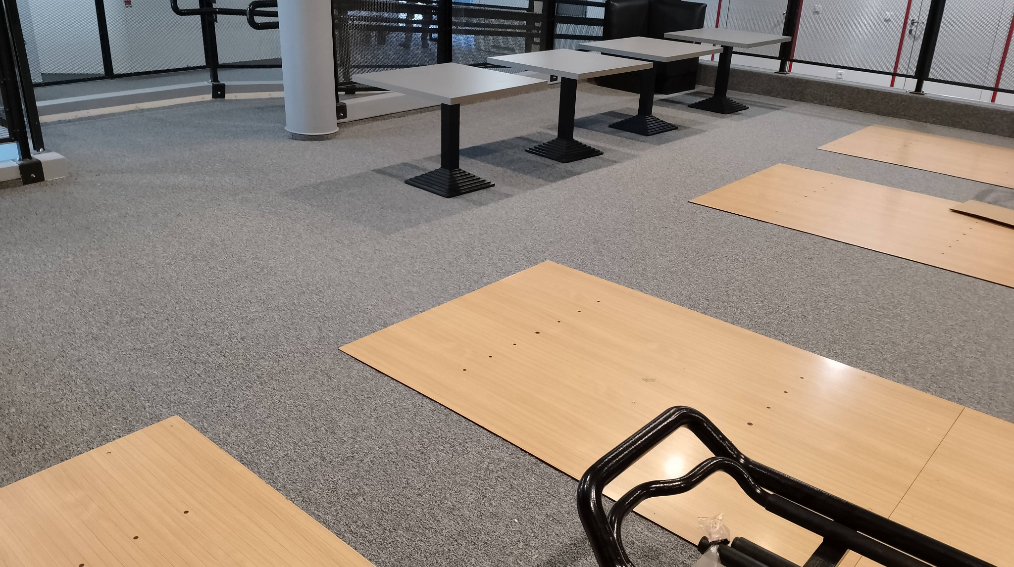 Montaż wykładziny dywanowej w przestrzeni biurowej, widoczne płyty wykładzinowe przed położeniem, w tle stoliki i krzesła, czarny wózek transportowy w rogu.