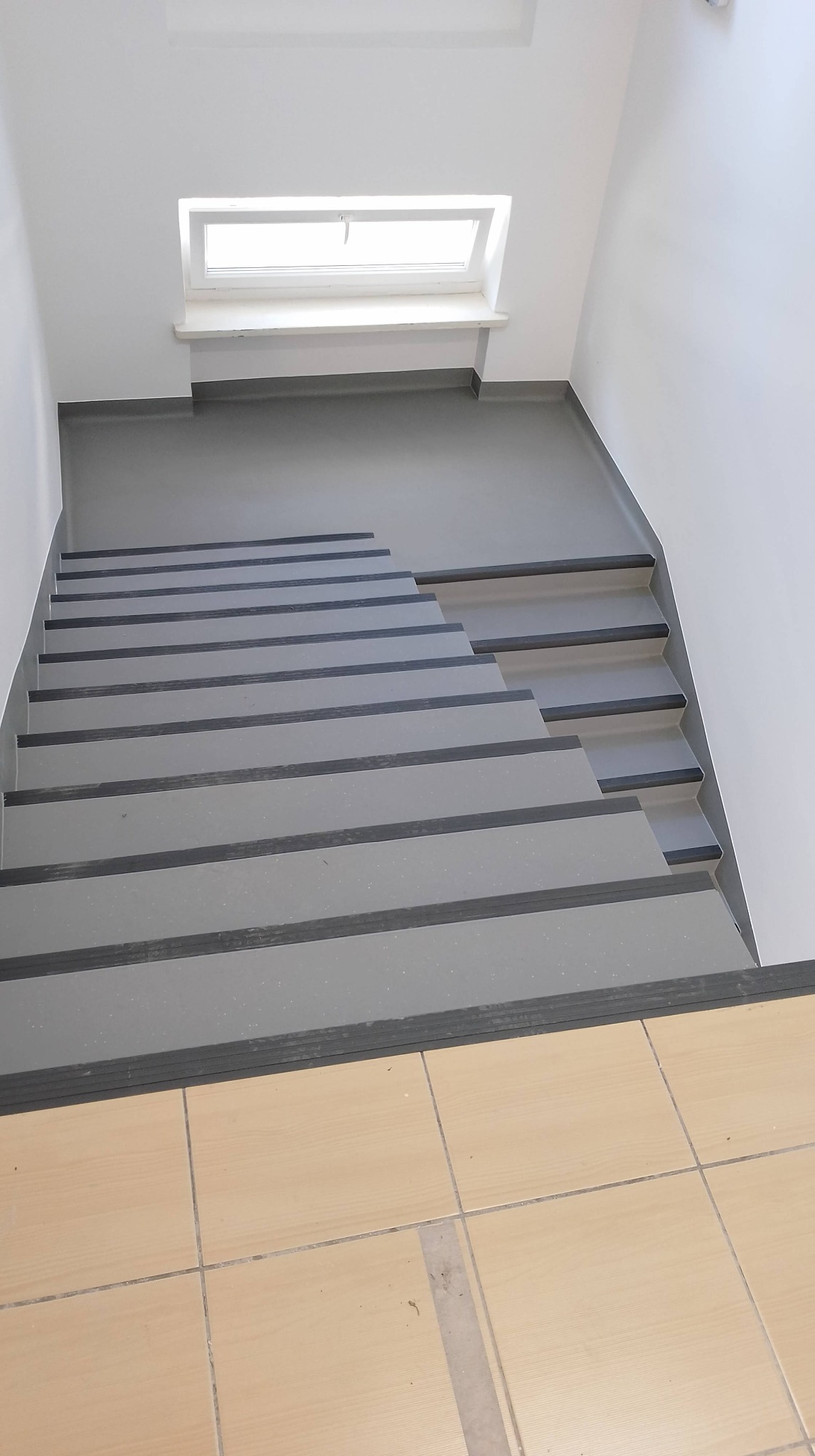 Schody pokryte szarą wykładziną z czarnymi pasami antypoślizgowymi, widok z góry na jasne płytki podłogowe.
