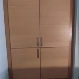 Wbudowana szafa z drewnianymi frontami i metalowymi uchwytami, umieszczona we wnęce ściany, z kratką wentylacyjną nad górną krawędzią.