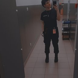 Abracadabra cleaning - Osoba w czarnym uniformie, z identyfikatorem i krzyżykiem na szyi, robi selfie w lustrze w korytarzu z płytkami. Widoczny fragment wózka z akcesoriami.