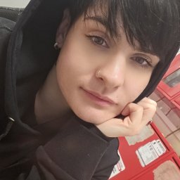 Abracadabra cleaning - Portret kobiety z krótkimi, ciemnymi włosami, w czarnej bluzie z kapturem, opierającej brodę na dłoni, z kolczykiem w nosie, na tle czerwonych szafek.