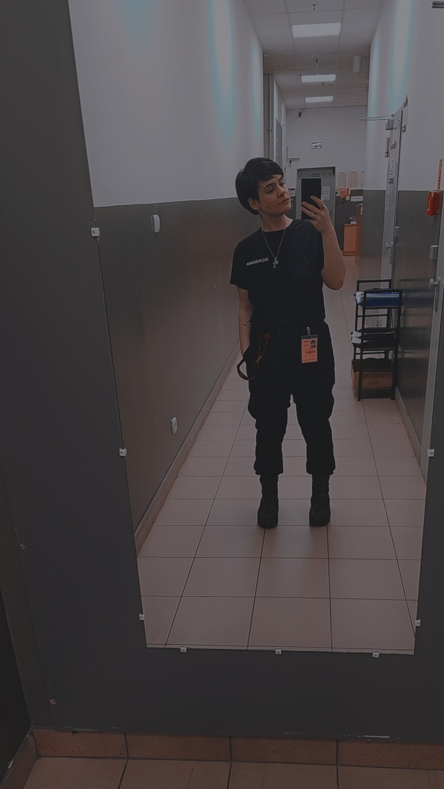 Osoba w czarnym uniformie, z identyfikatorem i krzyżykiem na szyi, robi selfie w lustrze w korytarzu z płytkami. Widoczny fragment wózka z akcesoriami.