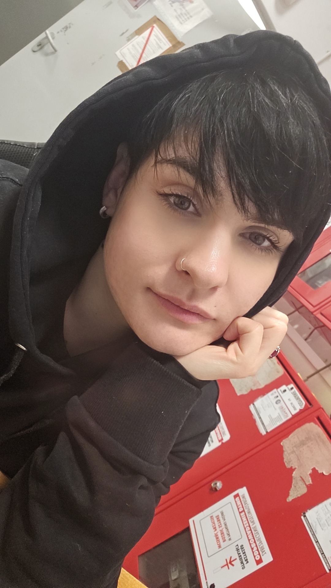 Portret kobiety z krótkimi, ciemnymi włosami, w czarnej bluzie z kapturem, opierającej brodę na dłoni, z kolczykiem w nosie, na tle czerwonych szafek.