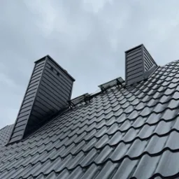 Nowoczesny, ciemnoszary dach z blachy trapezowej z dwoma kominami i zabezpieczeniami przeciwśniegowymi na tle pochmurnego nieba.