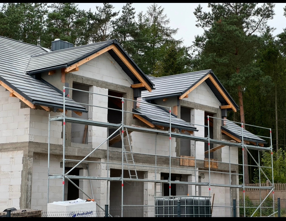 Budowa domu jednorodzinnego w stanie surowym otwartym z widocznym rusztowaniem i nowym dachem z szarej dachówki ceramicznej.