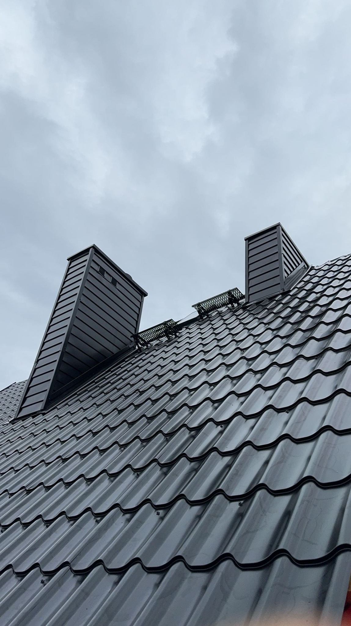 Nowoczesny, ciemnoszary dach z blachy trapezowej z dwoma kominami i zabezpieczeniami przeciwśniegowymi na tle pochmurnego nieba.