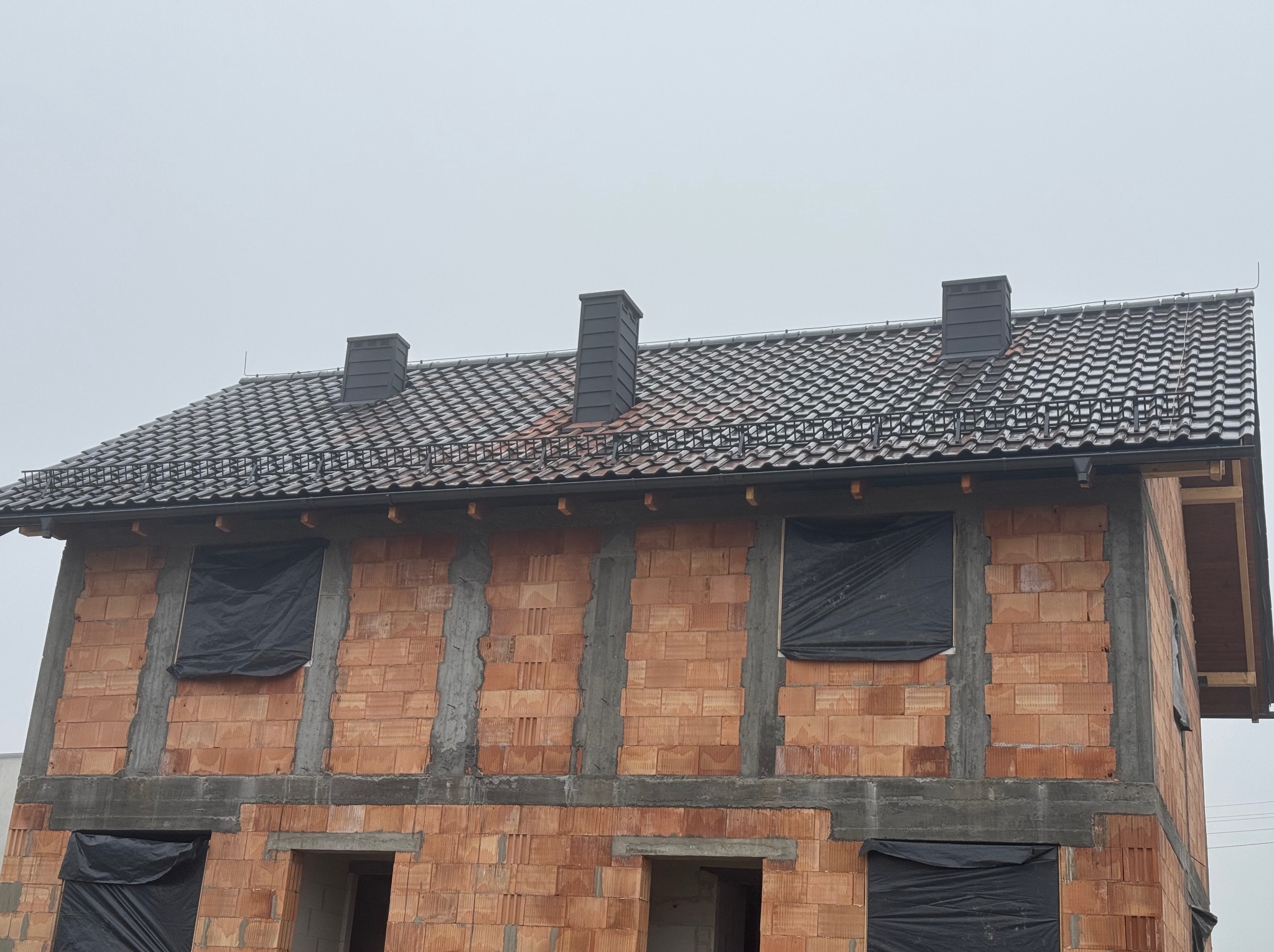 Surowy budynek w trakcie budowy z nowym dachem z ciemnej dachówki ceramicznej i trzema kominami, okna zabezpieczone czarną folią.