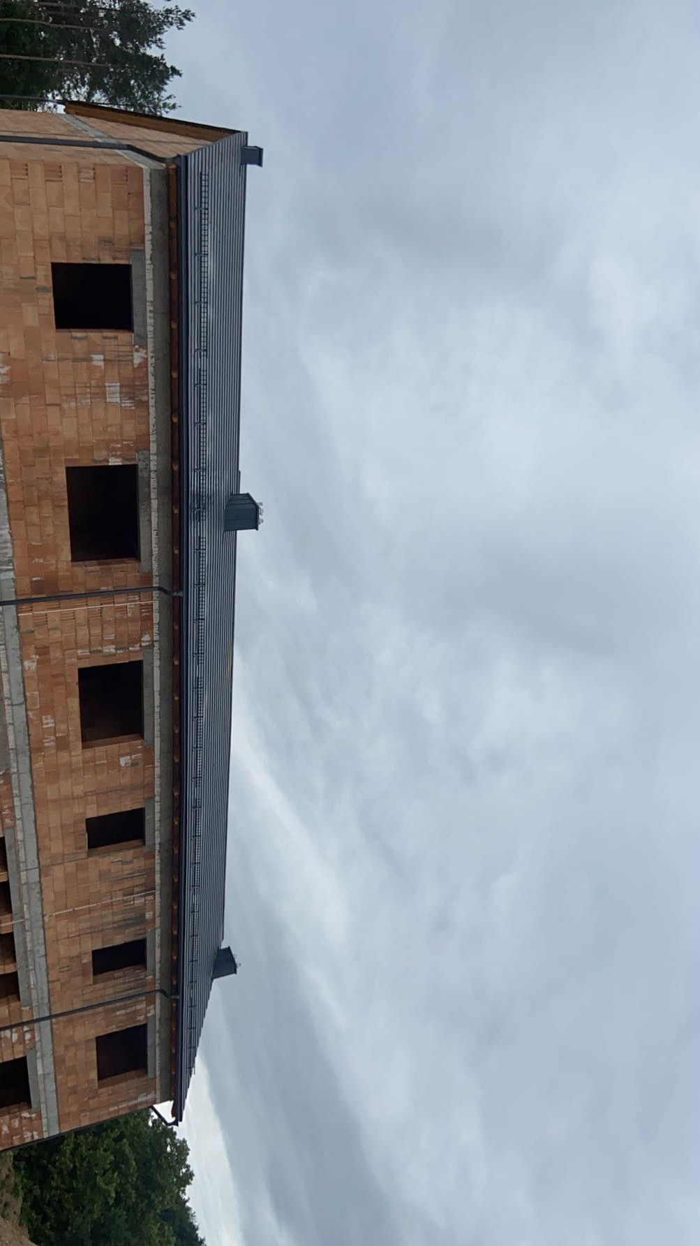 Wznoszony budynek z cegły z zamontowanym, szarym dachem blacharskim i rynnami, widok z dołu na tle pochmurnego nieba.