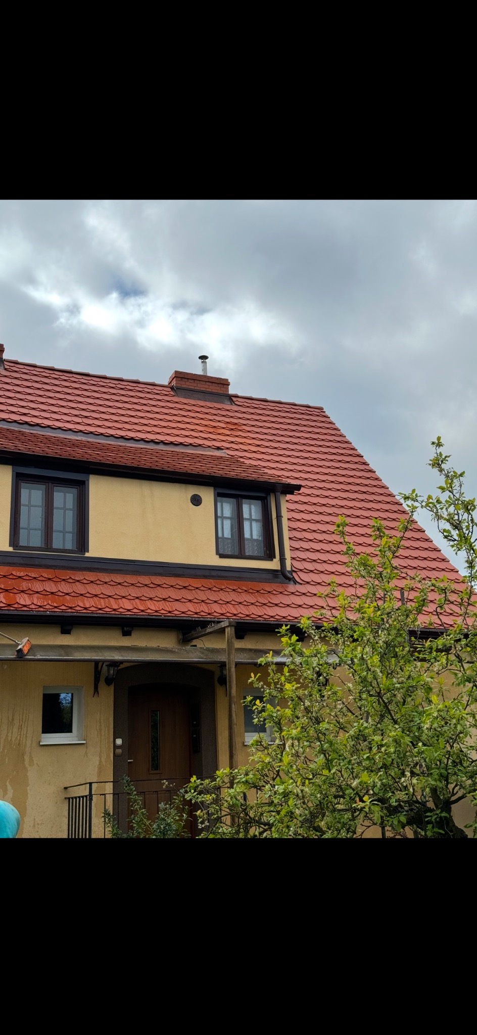 Dom jednorodzinny z nowym, czerwonym dachem z dachówki ceramicznej, widok z dołu na fragment fasady i dach z kominem na tle pochmurnego nieba.