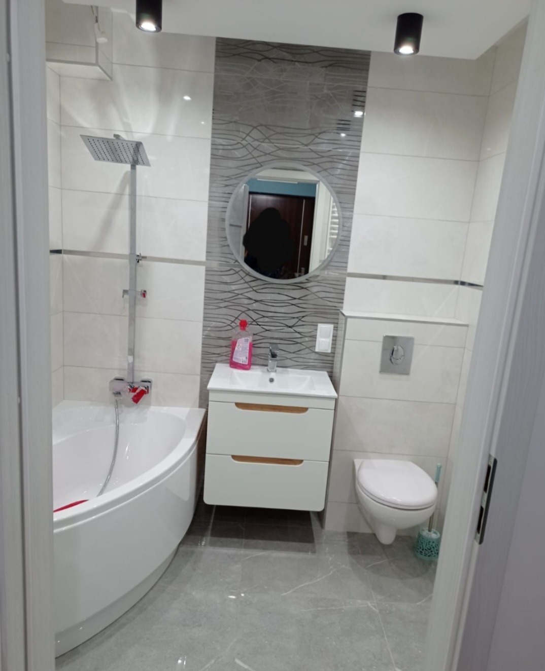 Nowoczesna łazienka z narożną wanną, umywalką z szafką w kolorze białym z drewnianymi akcentami, toaletą i szarą płytką dekoracyjną na ścianie za okrągłym lustrem.