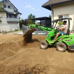 Ogrody Zielona Sowa - Praca ziemne w ogrodzie: operator na zielonej miniładowarce Avant 528 rozsypuje ziemię, przygotowując teren pod trawnik. W tle domy i pergola.