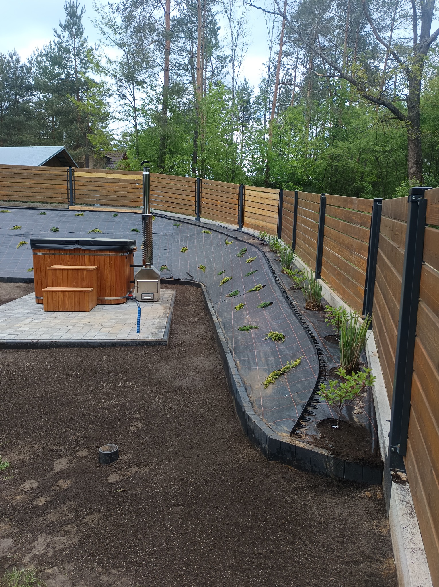 Fragment ogrodu w trakcie tworzenia: drewniana balia, ziemia przygotowana pod nasadzenia, agrowłóknina z rozsadami, drewniany płot z metalowymi słupkami.