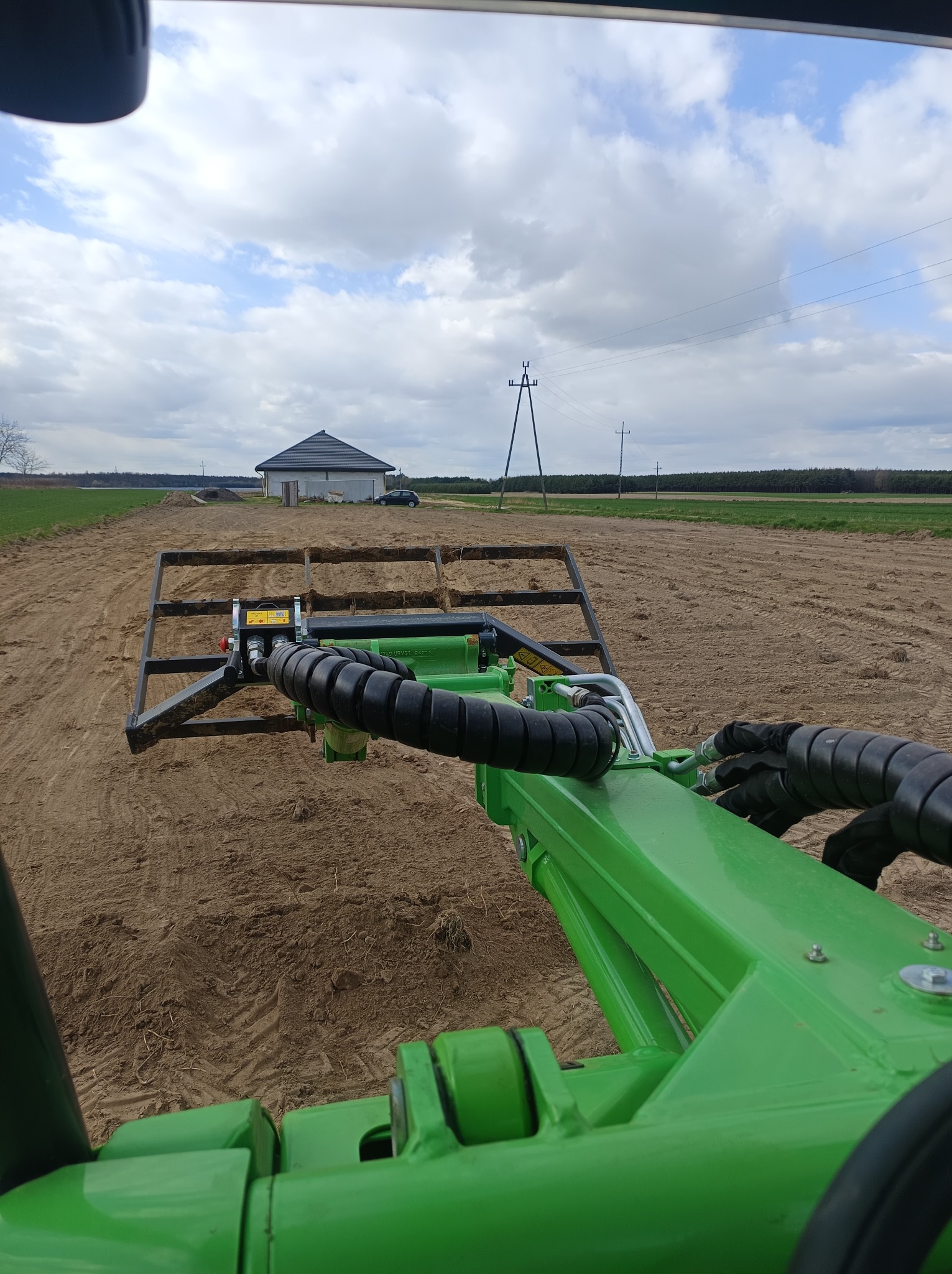 Widok z kabiny traktora na przygotowywaną glebę pod trawnik. W tle dom i pochmurne niebo. Specyficzny kąt ujęcia z wnętrza maszyny.