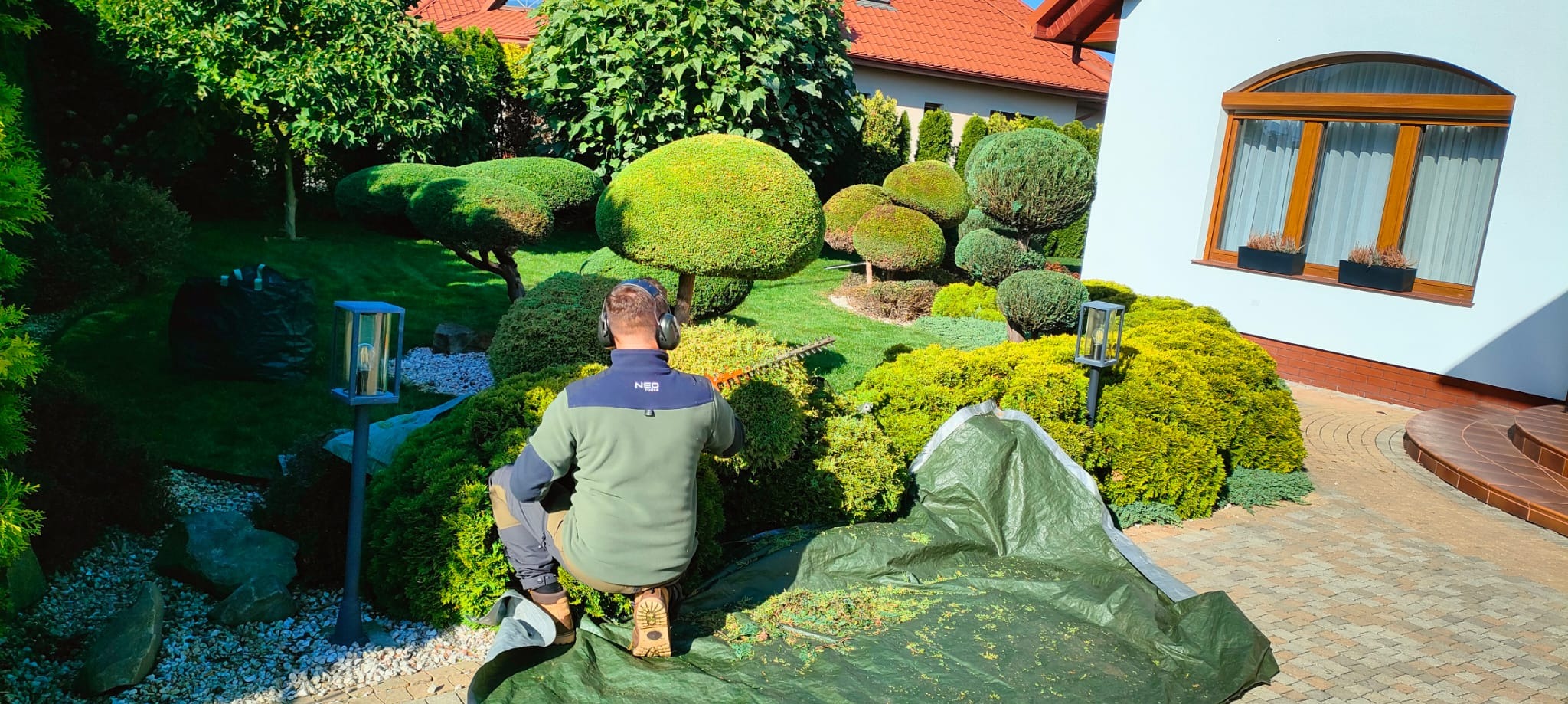 Ogrodnik w słuchawkach, z tyłu, formuje krzewy nożycami elektrycznymi w zadbanym ogrodzie z lampami i kostką brukową. Część obciętych gałęzi leży na plandece.