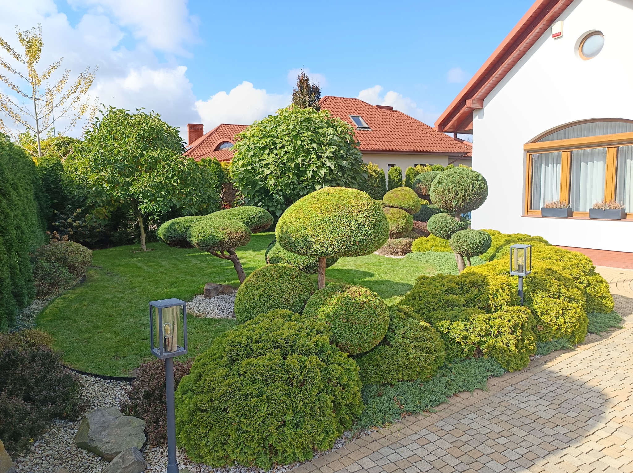 Ogród z równo przystrzyżonymi krzewami w formie kul i grzybów, trawnik, oświetlenie ogrodowe i kostka brukowa na tle domu z czerwonym dachem.
