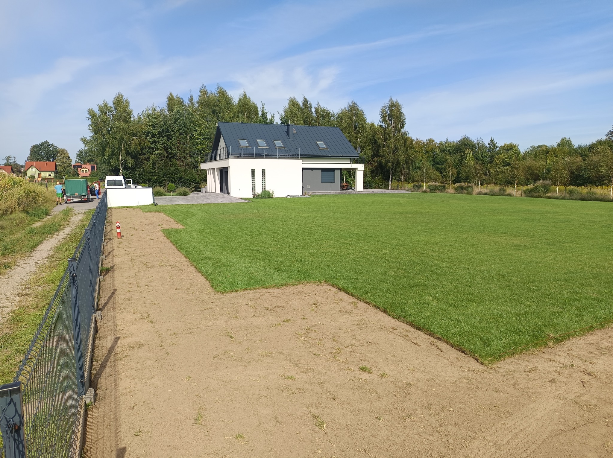 Nowo założony trawnik z widocznymi fragmentami przygotowanej ziemi, nowoczesny dom w tle i pracownicy w oddali, słoneczny dzień.