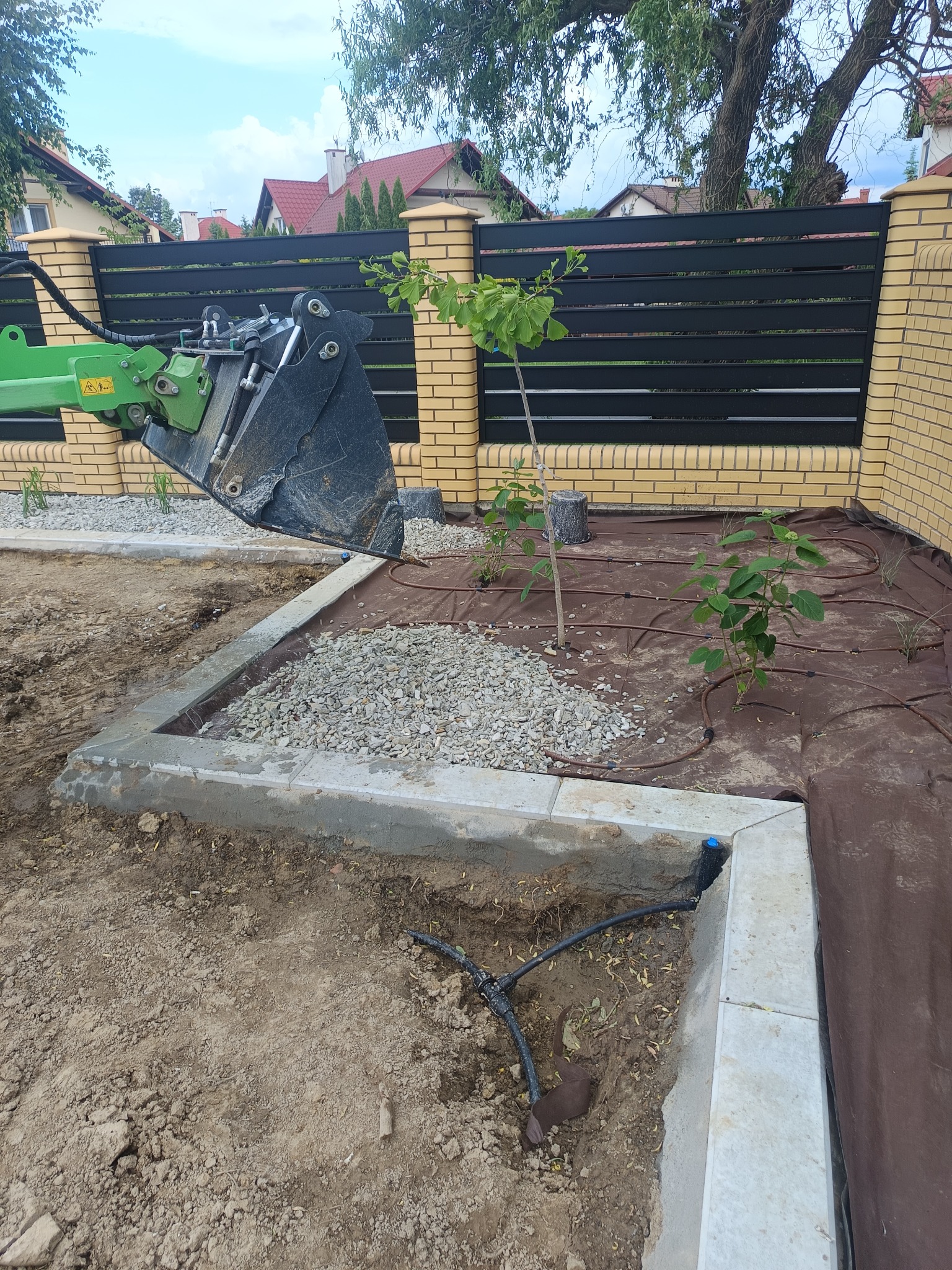 Prace ziemne w ogrodzie: koparka z łyżką nasypuje żwir na przygotowane rabaty z systemem nawadniania, w tle ogrodzenie z cegły i czarnych paneli.