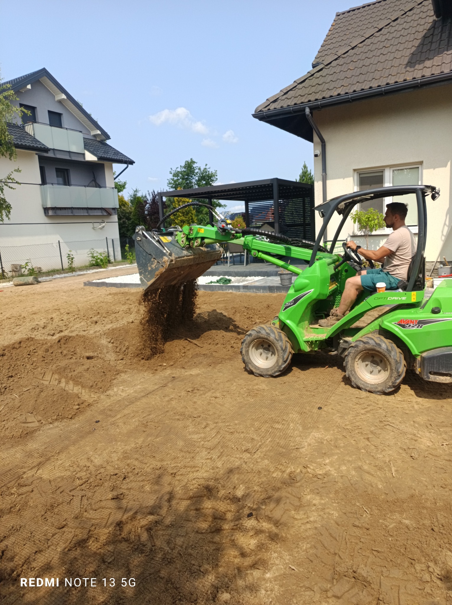 Praca ziemne w ogrodzie: operator na zielonej miniładowarce Avant 528 rozsypuje ziemię, przygotowując teren pod trawnik. W tle domy i pergola.