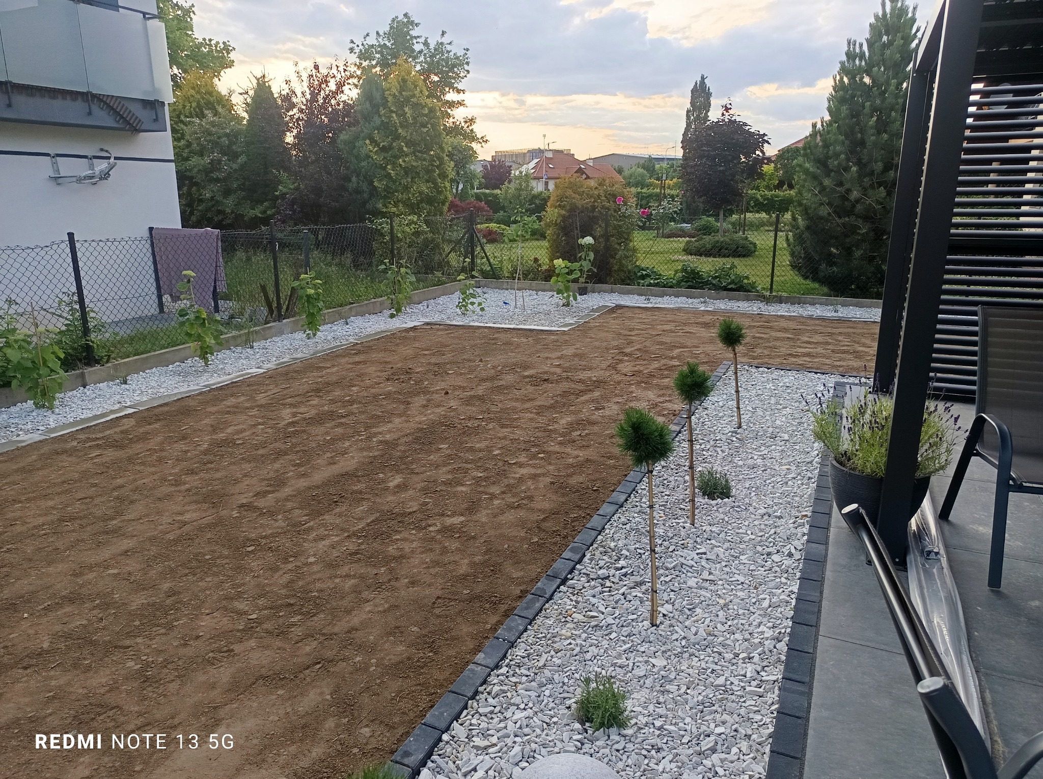 Przygotowany teren pod trawnik z obrzeżami z kostki i białego kamienia, ozdobne drzewka, widok z tarasu. W tle zabudowa jednorodzinna i zieleń.