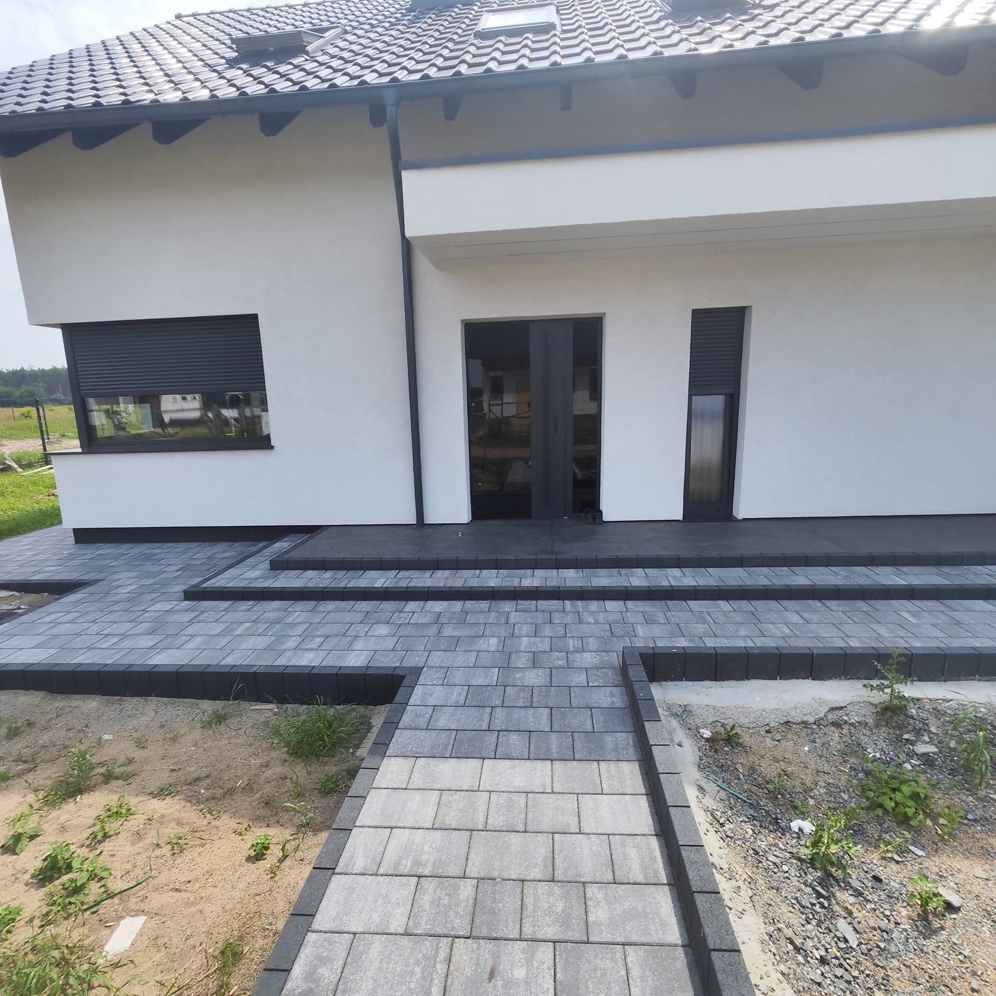 Nowoczesny podjazd z szarej kostki brukowej prowadzący do wejścia do domu z ciemnymi oknami i drzwiami.