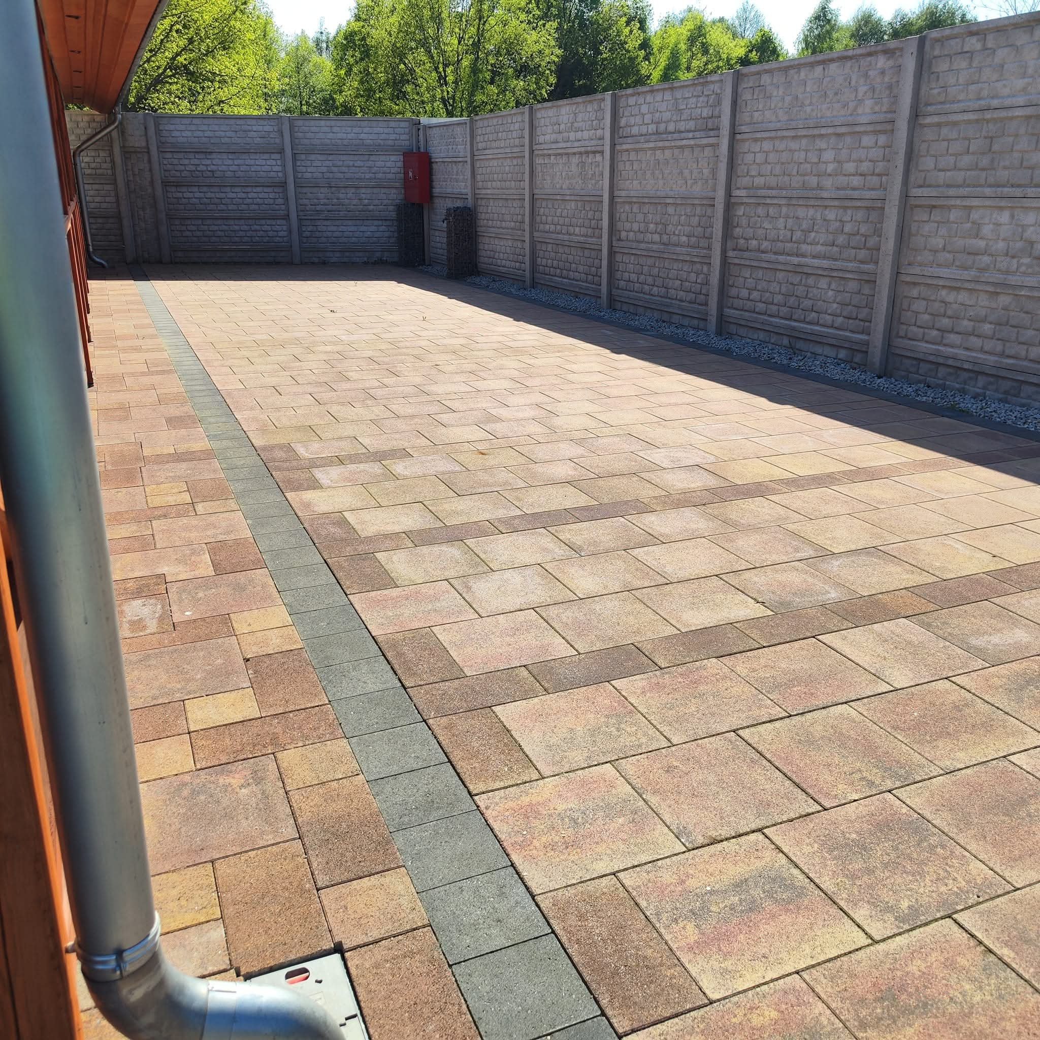 Ułożona kostka brukowa w odcieniach brązu i beżu, obrzeże z szarej kostki, w tle betonowy płot z elementami dekoracyjnymi, widoczna rynna spustowa z boku.