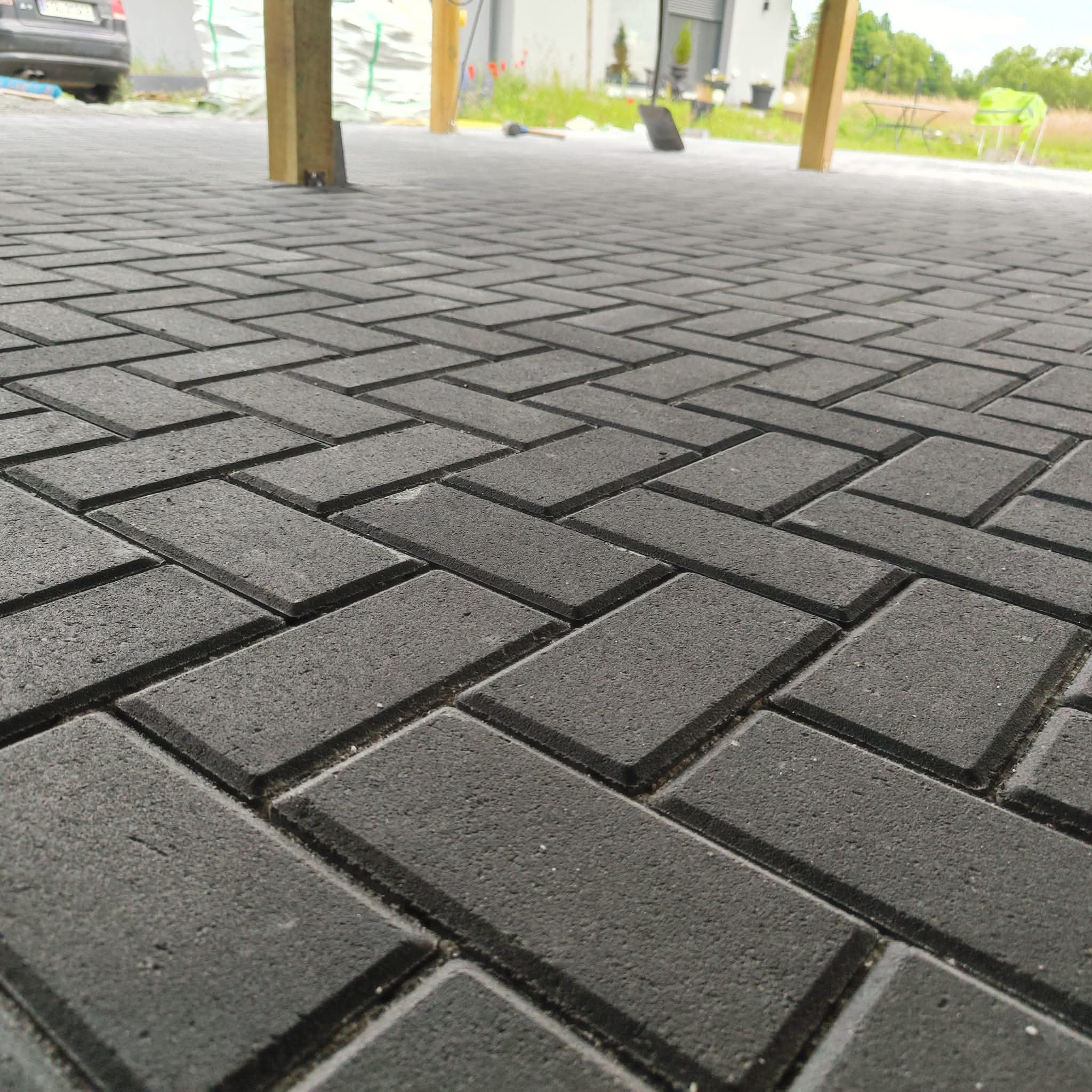 Nowa, ciemnoszara nawierzchnia z kostki brukowej ułożonej w jodełkę, widok z niskiej perspektywy z elementami drewnianej konstrukcji w tle.
