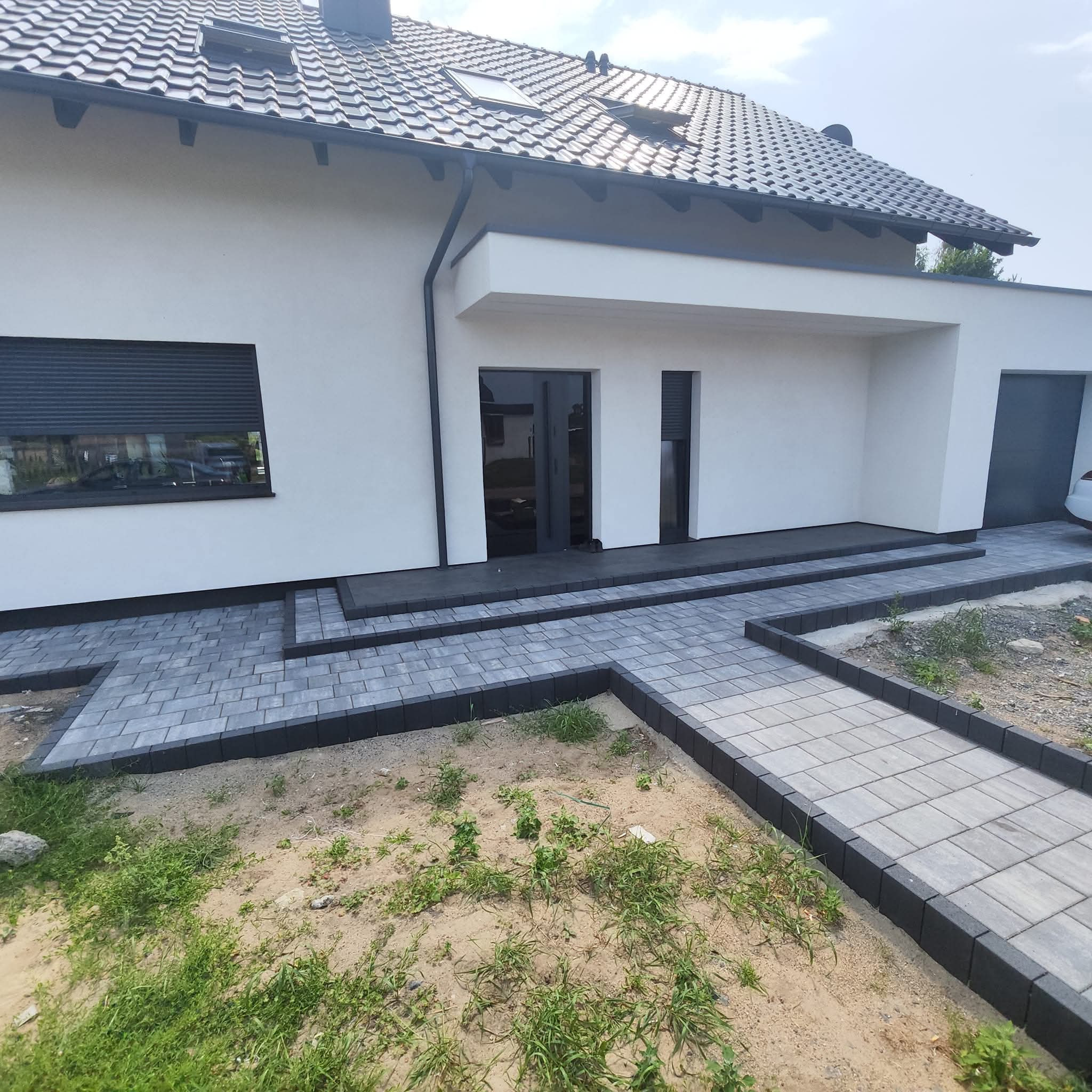 Nowoczesny dom z szarą kostką brukową prowadzącą do wejścia, widoczny fragment ogrodu z młodą trawą i piaskiem.