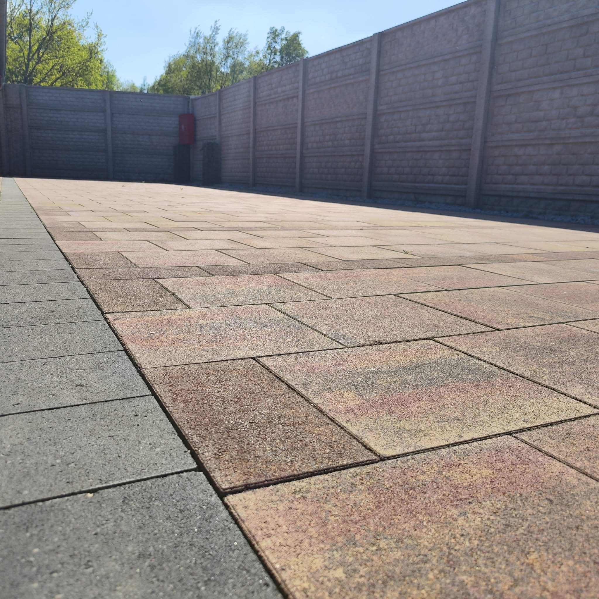 Ułożona kostka brukowa w odcieniach brązu i szarości, ograniczona betonowym ogrodzeniem panelowym z widocznym zielonym drzewostanem w tle.