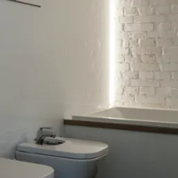 Nowoczesna łazienka z białą armaturą, bidetem, wanną obudowaną drewnianym elementem i ścianą z białej cegły podświetloną pionowym paskiem LED.