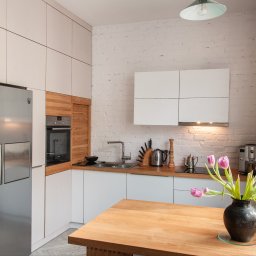 Arser Wood Polska s.c. Sławomir Brzózka, Tomasz Matosz - Jasna kuchnia z białymi szafkami, drewnianymi akcentami i ceglaną ścianą, uzupełniona o drewniany stół z tulipanami w wazonie i misą owoców.