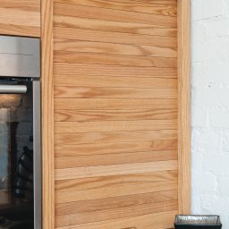 Arser Wood Polska s.c. Sławomir Brzózka, Tomasz Matosz - Drewniana roleta meblowa z poziomo ułożonych listew, zabudowa kuchenna z piekarnikiem, obok ściana z białej cegły