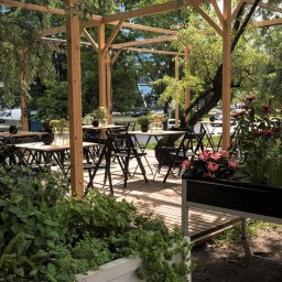 Arser Wood Polska s.c. Sławomir Brzózka, Tomasz Matosz - Drewniana pergola z zadaszeniem, podłogą z desek i czarnymi krzesłami, otoczona zielenią i kwiatami, tworząca letnią restaurację na świeżym powietrzu.