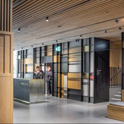 Arser Wood Polska s.c. Sławomir Brzózka, Tomasz Matosz - Wnętrze hotelu Nobu w Warszawie: recepcja z metalowym frontem i logo, drewniana boazeria na ścianach i suficie, betonowe elementy dekoracyjne, pracownicy hotelu w tle.