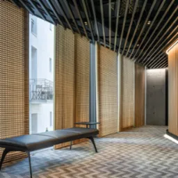 Nowoczesne wnętrze z drewnianą ażurową ścianą, czarną ławką i wzorzystą wykładziną podłogową, widok na balkon przez okno.