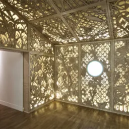 Wnętrze altany z ażurowymi ścianami i sufitem z drewna, podświetlonymi od tyłu, tworzącymi wzór roślinny; okrągłe okno w jednej ze ścian, drewniana podłoga.