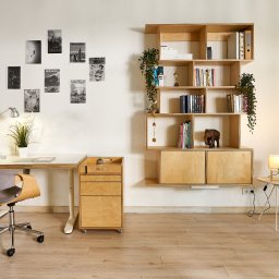 Arser Wood Polska s.c. Sławomir Brzózka, Tomasz Matosz - Jasne biuro z drewnianym biurkiem, krzesłem na kółkach, kontenerkiem z szufladami i wiszącą półką na książki z roślinami, uzupełnione lampką na biurku i stoliku nocnym oraz plakatami na ścianie.