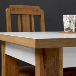 Biały blat stołu z drewnianym obramowaniem i nogami z surowego drewna, w tle krzesło z drewna i kubek z wzorem na podkładce.