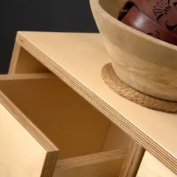 Szafka z jasnej sklejki z wysuwanymi szufladami, na blacie drewniana misa z brązowym paskiem skórzanym z wycinanym wzorem.