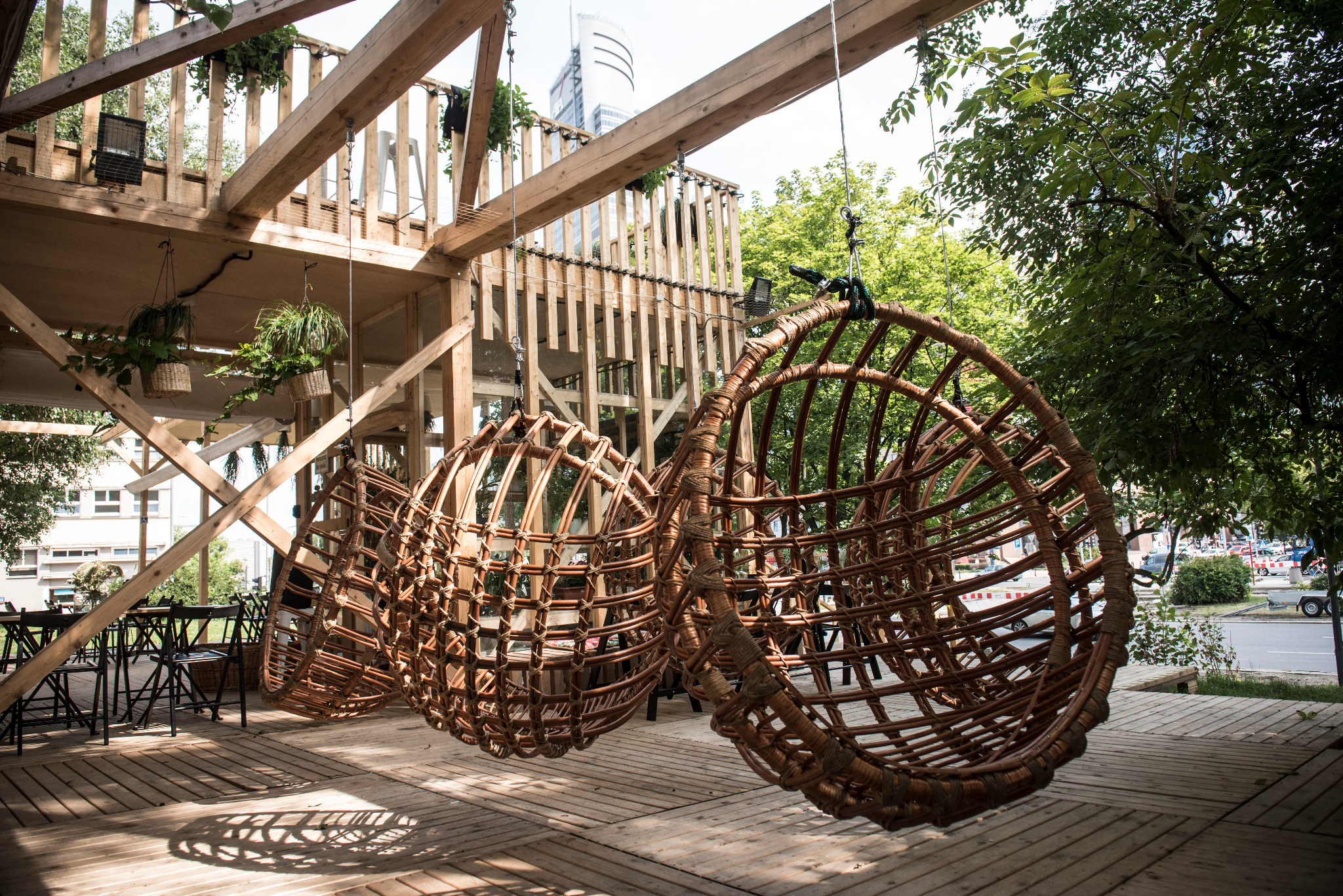Trzy wiszące, rattanowe fotele typu kokon na drewnianej konstrukcji z widocznym miejskim krajobrazem w tle.