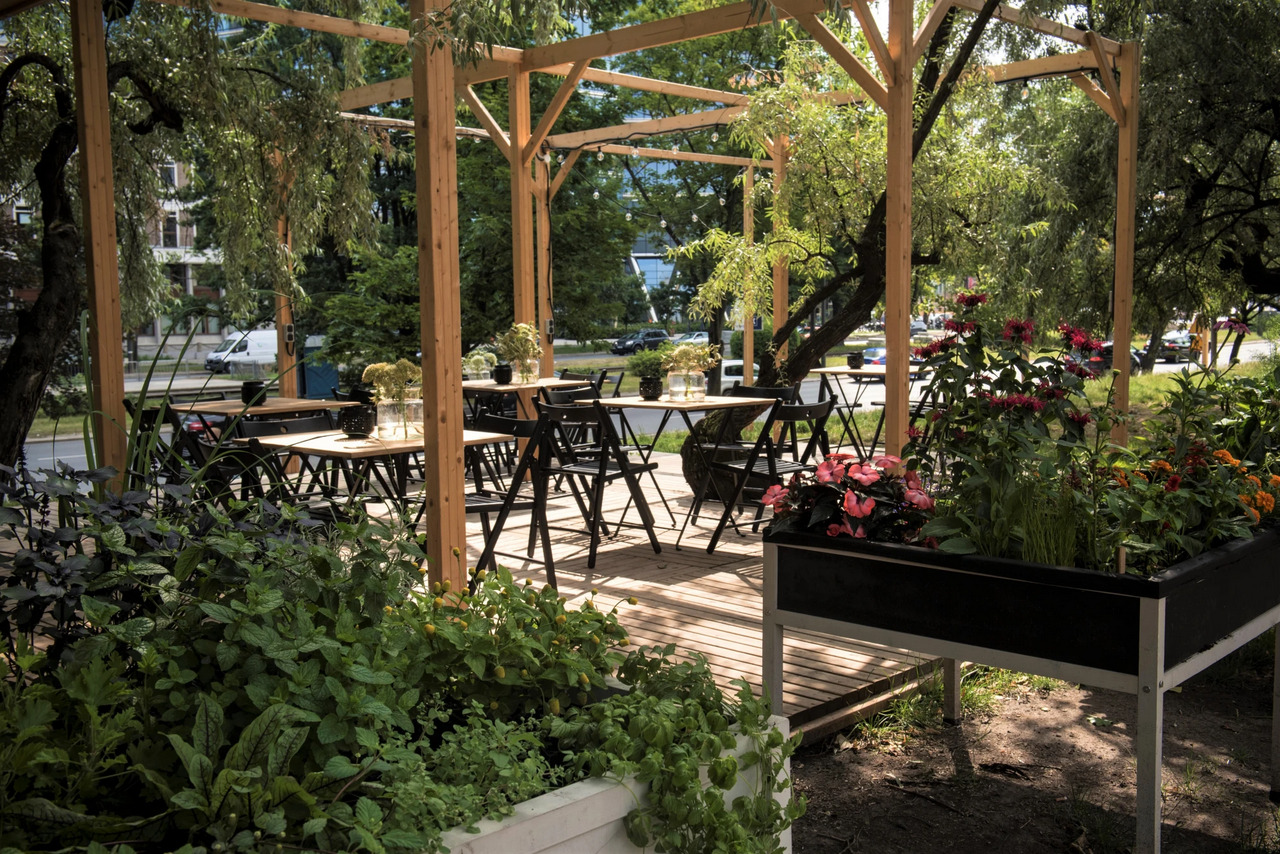 Drewniana pergola z zadaszeniem, podłogą z desek i czarnymi krzesłami, otoczona zielenią i kwiatami, tworząca letnią restaurację na świeżym powietrzu.