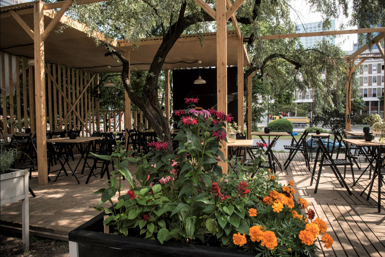Drewniana konstrukcja z zadaszeniem, tworząca otwartą przestrzeń restauracyjną z drewnianą podłogą, stolikami i krzesłami, z roślinami w donicach na pierwszym planie i drzewem wrastającym...