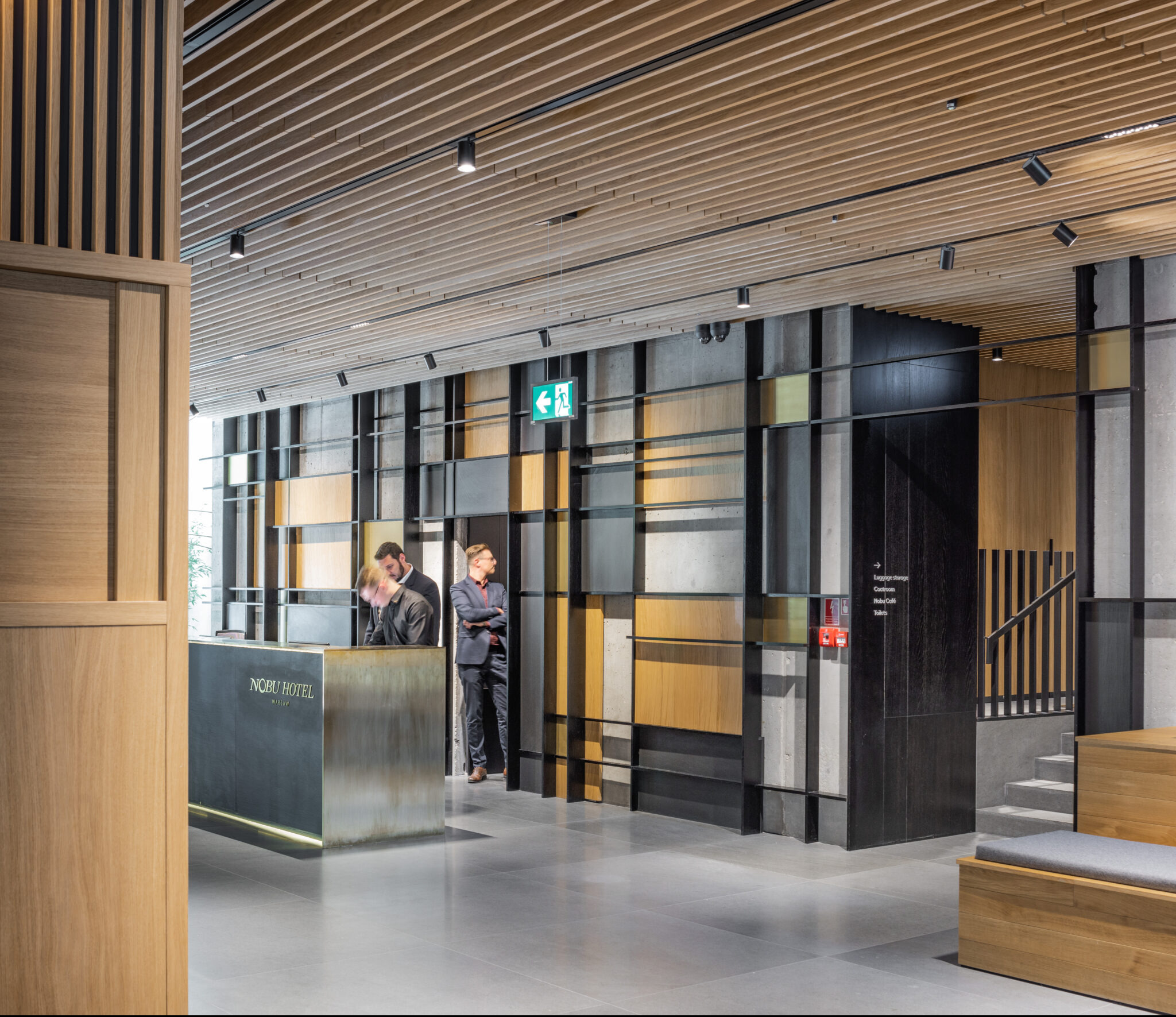 Wnętrze hotelu Nobu w Warszawie: recepcja z metalowym frontem i logo, drewniana boazeria na ścianach i suficie, betonowe elementy dekoracyjne, pracownicy hotelu w tle.