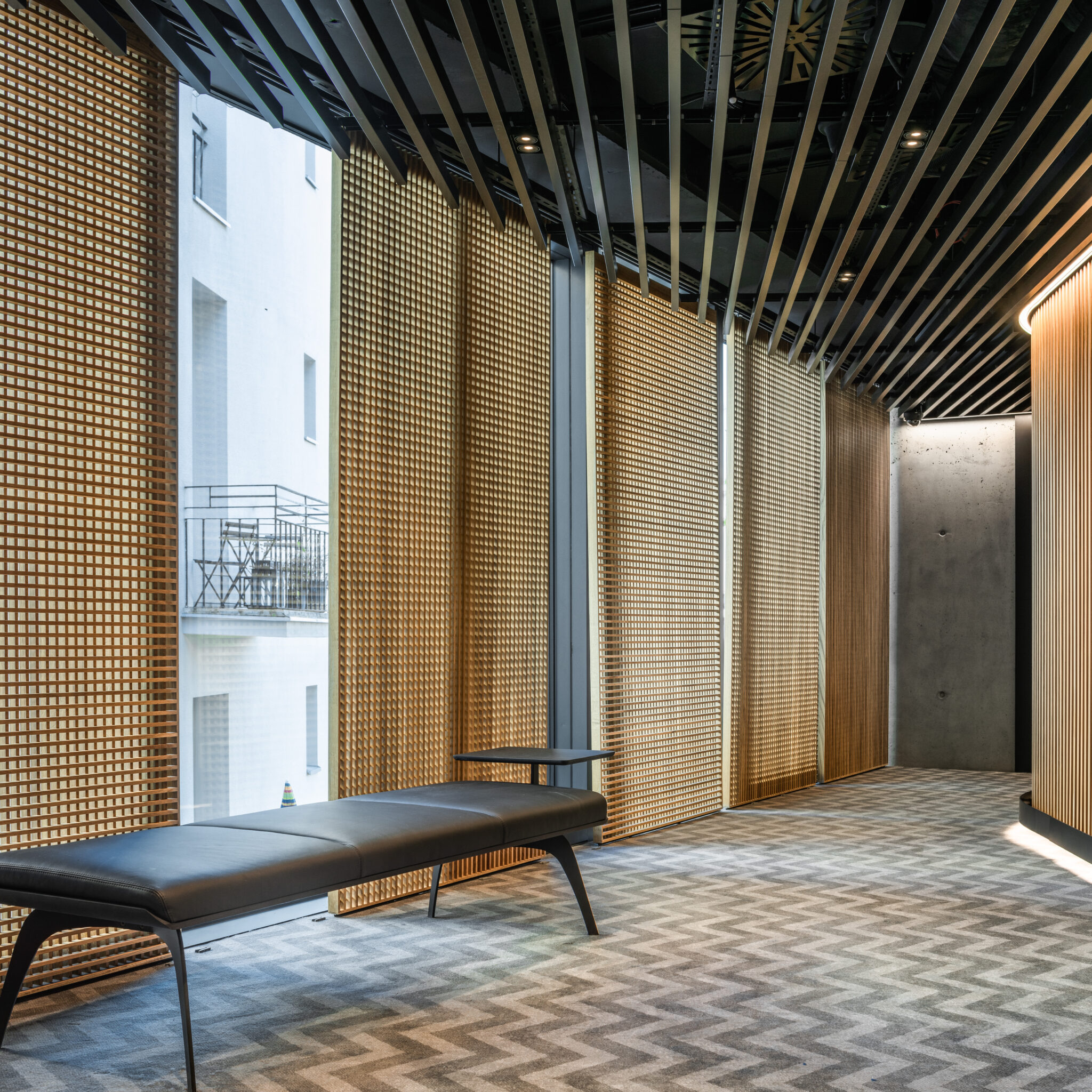 Nowoczesne wnętrze z drewnianą ażurową ścianą, czarną ławką i wzorzystą wykładziną podłogową, widok na balkon przez okno.